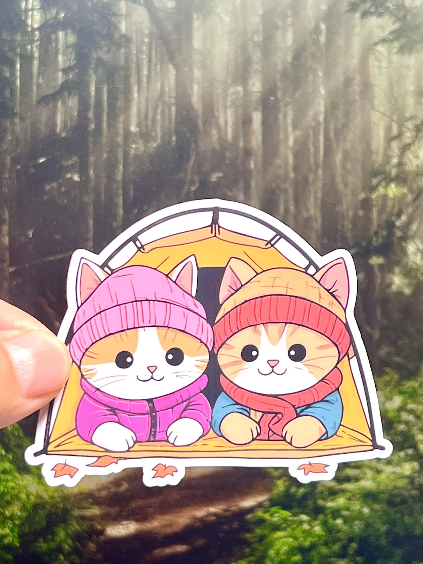 A sticker depicting two cartoon cats with pink hats and blue eyes, peeking out from a camping tent in a forest setting.