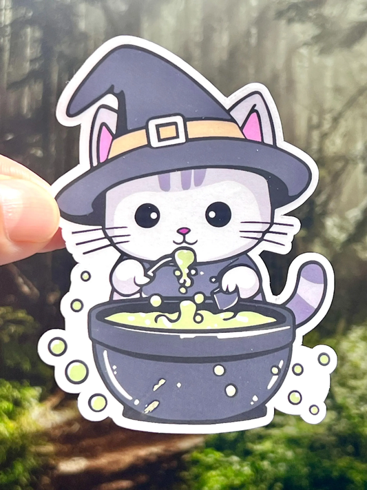 Sticker of a cat dressed like a witch with a black hat and standing behind a bubbling cauldron